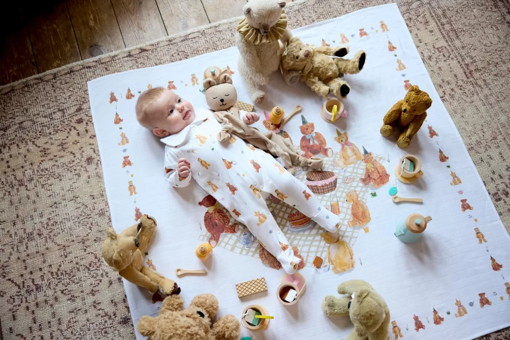 Baby wearing peter pan collared sleepsuit surrounded by a bear comforter, teddies and lying on a large muslin blanket. All items are themed as a teddy bears picnic